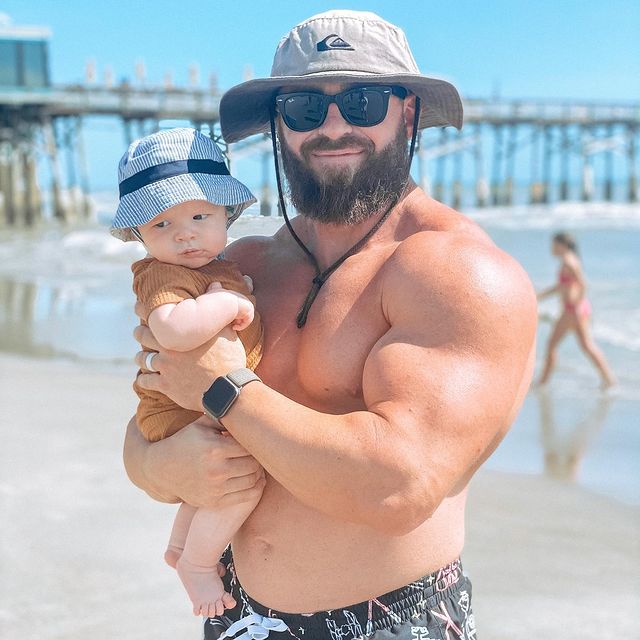 Tanned man in a hat and bathing suit holding a baby in a bathing suit and hat